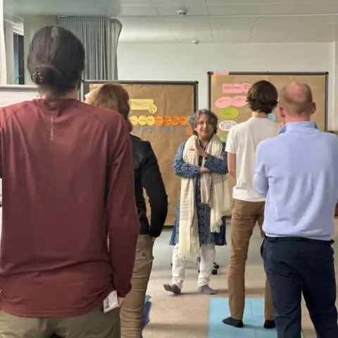 A group of people stand in a room, facing a woman who appears to be leading a discussion. 