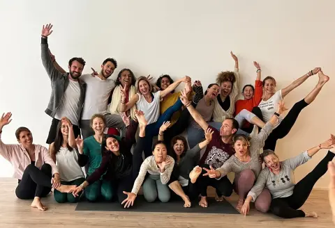 A joyful group of people pose energetically, with wide smiles and raised arms, in a yoga studio. 