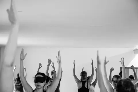 Black and white photo of a blindfolded meditation or yoga class where participants have their arms outstretched toward the ceiling.