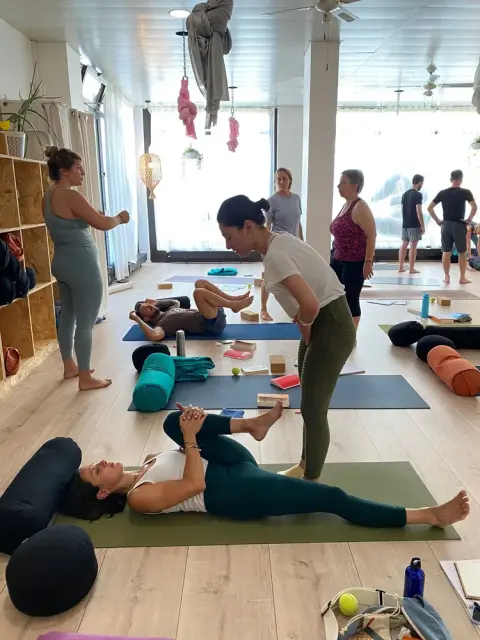 A yoga class with multiple people. A woman assists another lying on a mat, surrounded by props. The atmosphere is focused and serene.