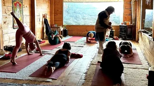 An instructor leads a small group yoga class in a rustic wooden cabin with a large window overlooking a forest.