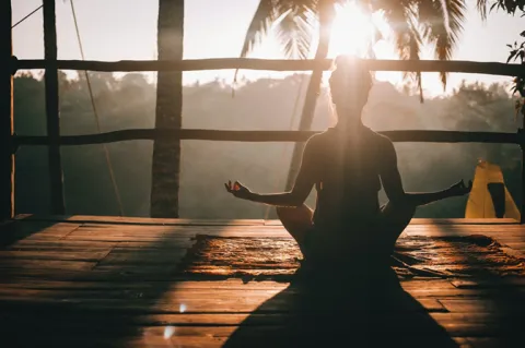 A silhouette of a person meditating in a lotus pose on a wooden deck during a tropical sunset.
