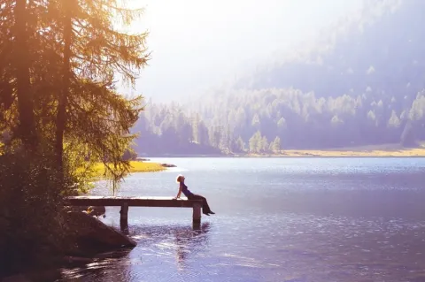 Silhouette of a person relaxing on a wooden dock overlooking a calm alpine lake surrounded by evergreen forests in golden sunlight.