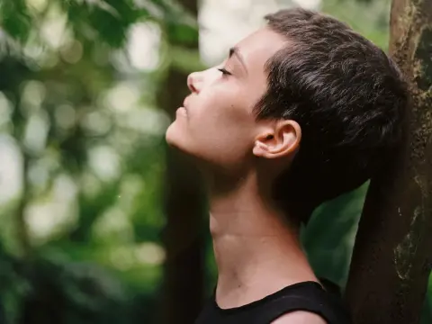 A peaceful side profile of a person meditating or breathing deeply in a sun-dappled woodland.