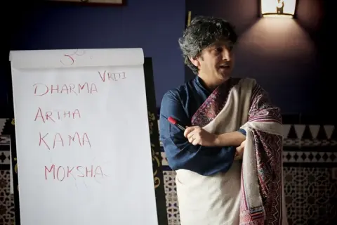 Sanjeev Bhanot stands by a flip chart listing Dharma, Artha, Kama, and Moksha during a philosophy lecture.