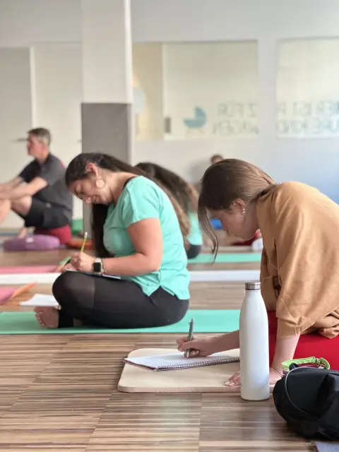 People sitting cross-legged on yoga mats in a calm studio, writing in notebooks. 