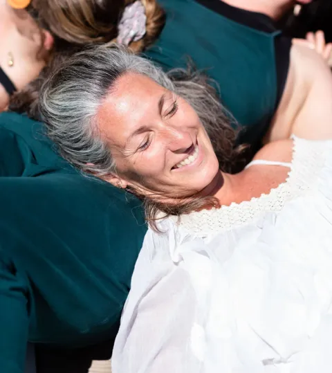 A close-up of a smiling woman with grey hair resting her head on a friend's back during an outdoor gathering.