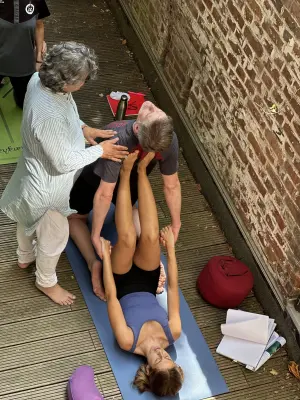 A yoga instructor provides a hands-on adjustment to a student performing an inverted pose on a mat against a brick wall.