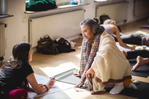 A woman and an older man sit on yoga mats in a sunlit room. He wears a robe and scarf, speaking with a thoughtful expression. 