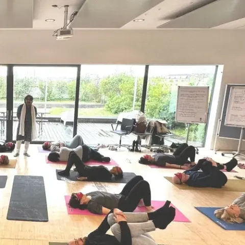A calm yoga class with participants lying on mats in a bright studio. An instructor stands beside large windows showing greenery outside.