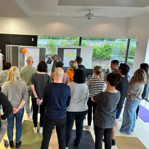 A group of people stands indoors facing two boards with sticky notes, while one person appears to be presenting at the front of the room.