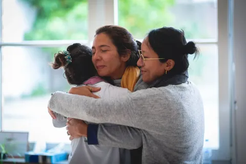 Three women of diverse backgrounds share a close, emotional group hug indoors near a window.