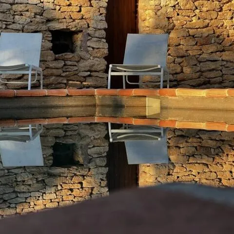 Two lounge chairs against a rustic stone wall, perfectly mirrored in the still water of a pool.