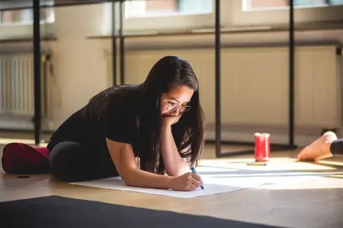 A young woman with glasses sits on a wooden floor, leaning over a large sheet of paper while drawing with a marker in a sunlit studio.