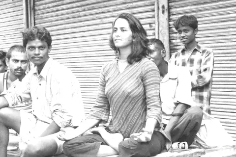 A black and white photo of a woman meditates in a lotus pose outdoors on a platform, surrounded by several men in a street setting.