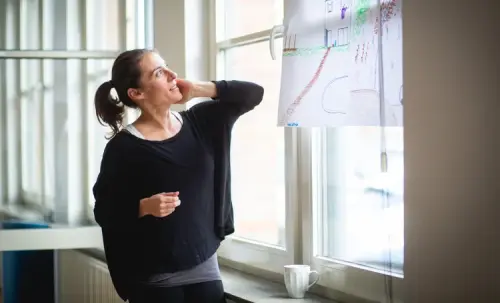 A smiling woman in a black sweater stands by a window, looking at a colorful sketch hanging on the glass in a bright studio.