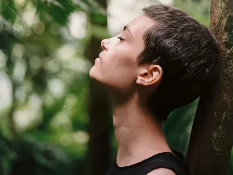 A profile view of a woman with short hair leaning against a tree in a lush forest with her eyes closed in meditation.