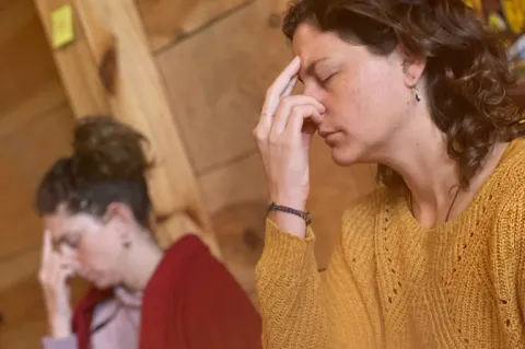 Two women practicing alternate nostril breathing meditation with eyes closed in a room with wood-paneled walls.