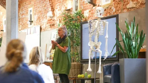 An instructor in a green dress stands next to a human skeleton model while teaching a class in a room with brick walls.