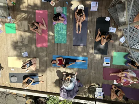 An overhead view of a group of people sitting and lying on colorful yoga mats on a wooden deck during a sunlit class.