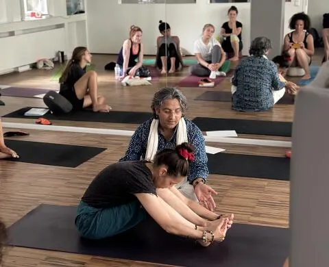 Man assisting a woman practicing Paschimottanasana stretch on a yoga mat with other students watching in the background.