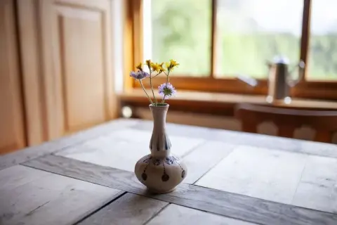 A small ceramic vase with delicate wildflowers sitting on a rustic wooden table by a bright window.
