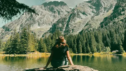 A woman wearing a hat sitting on a rock looking at a mountain lake surrounded by pine trees.
