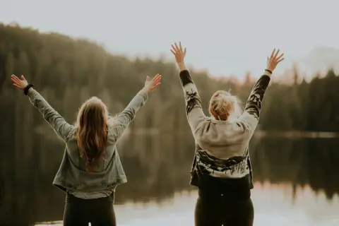 Two women standing back-to-back by a lake raising their arms up in celebration.